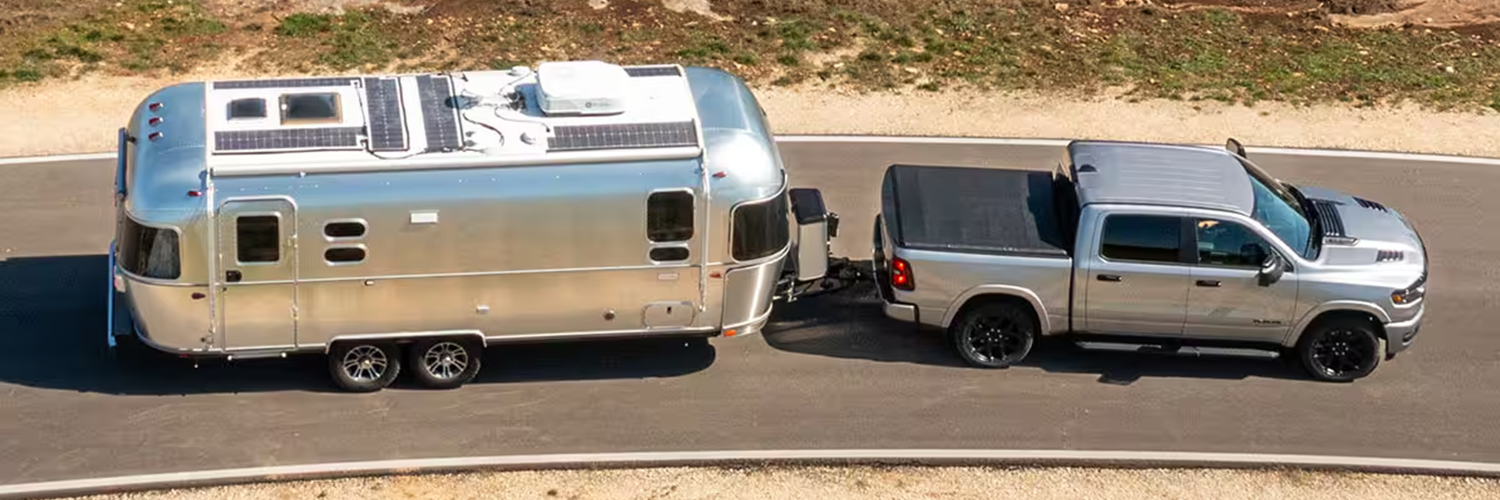 A raised angle displaying a silver 2025 Ram 1500 Laramie Crew Cab with tonneau cover, towing a large travel trailer as it's driven down a highway.
